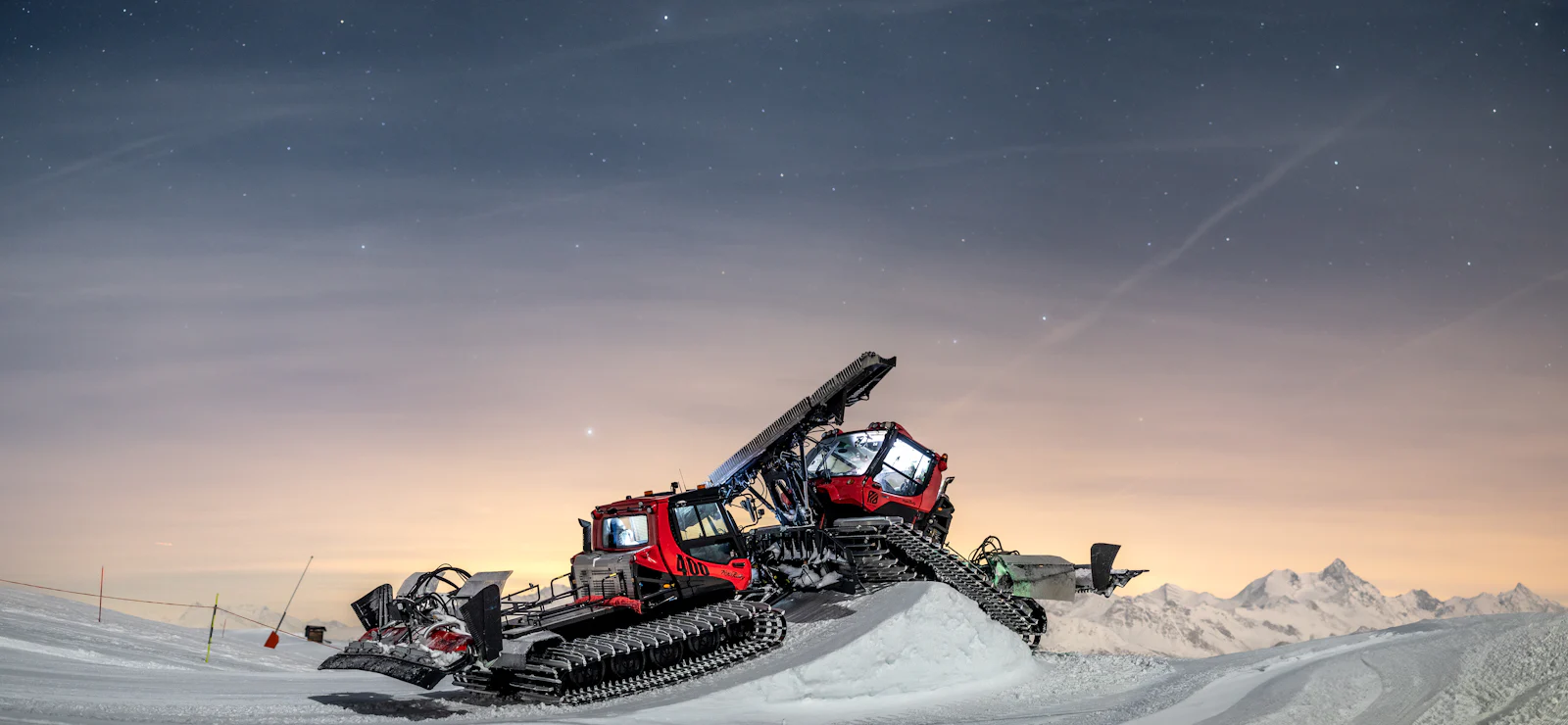 Two red snow groomers rebuilding a ski slope at dusk under a starry alpine sky